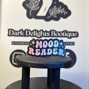 May include: A black and white sticker with the text "MOOD READER" in rainbow colors. The sticker is on a black wooden stand. The sticker is a handmade item from Dark Delights Boutique.