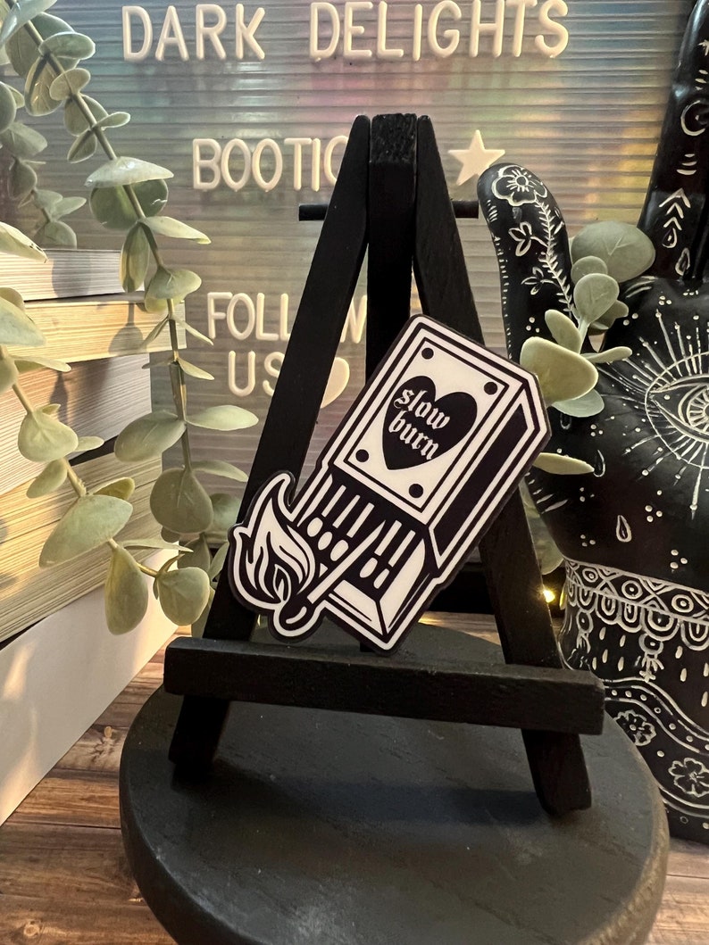 May include: Black and white sticker of a matchbox with a flame and the text "slow burn" on a small black easel.