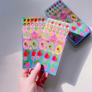 May include: Holographic sticker sheets with colourful designs. The stickers feature strawberries, cakes, hearts, and stars. The sheets have a rainbow sheen and are held in a hand. The stickers are on a white surface.