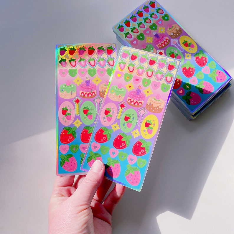 May include: Holographic sticker sheets with colourful designs. The stickers feature strawberries, cakes, hearts, and stars. The sheets have a rainbow sheen and are held in a hand. The stickers are on a white surface.