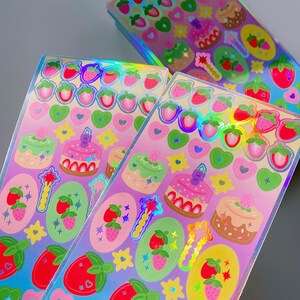 May include: Close-up of sticker sheets with a holographic finish. The stickers feature strawberries, cakes, hearts, stars, and magic wands. The colour palette includes pink, green, red, and yellow.