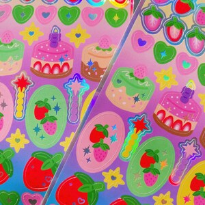May include: A sheet of colourful stickers featuring cakes, strawberries, hearts, stars, and magic wands. The stickers have a holographic, iridescent finish and are set against a gradient pink and purple background. Ideal for scrapbooking or crafts.