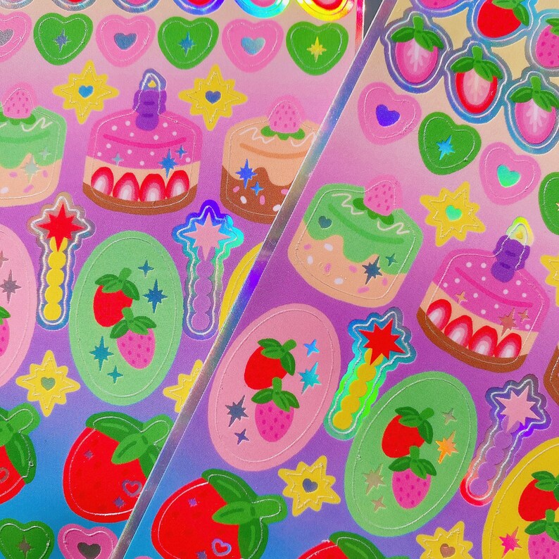 May include: A sheet of colourful stickers featuring cakes, strawberries, hearts, stars, and magic wands. The stickers have a holographic, iridescent finish and are set against a gradient pink and purple background. Ideal for scrapbooking or crafts.