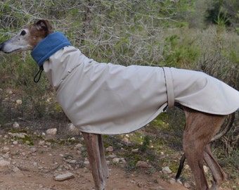 Può includere: Un cappotto beige per cani con fodera in pile blu. Il cappotto è progettato per un cane con gambe lunghe e una corporatura sottile. Il cane indossa il cappotto all'aperto in un ambiente naturale.