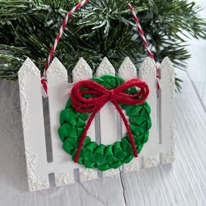 May include: A white picket fence Christmas ornament with a green ribbon wreath and a red bow. The ornament is decorated with a red and white string for hanging. The background features evergreen branches.