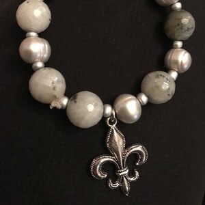 May include: A silver bracelet with alternating grey and white beads, featuring a silver fleur-de-lis charm.