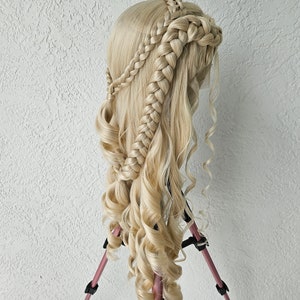 May include: A blonde wig with long, curly hair styled in a braided crown. The wig is mounted on a pink tripod.