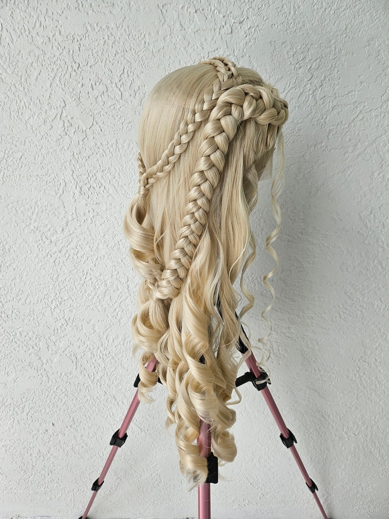 May include: A blonde wig with long, curly hair styled in a braided crown. The wig is mounted on a pink tripod.