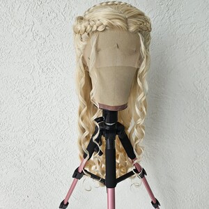 May include: A blonde wig with long, loose curls and a braided crown. The wig is displayed on a pink tripod.