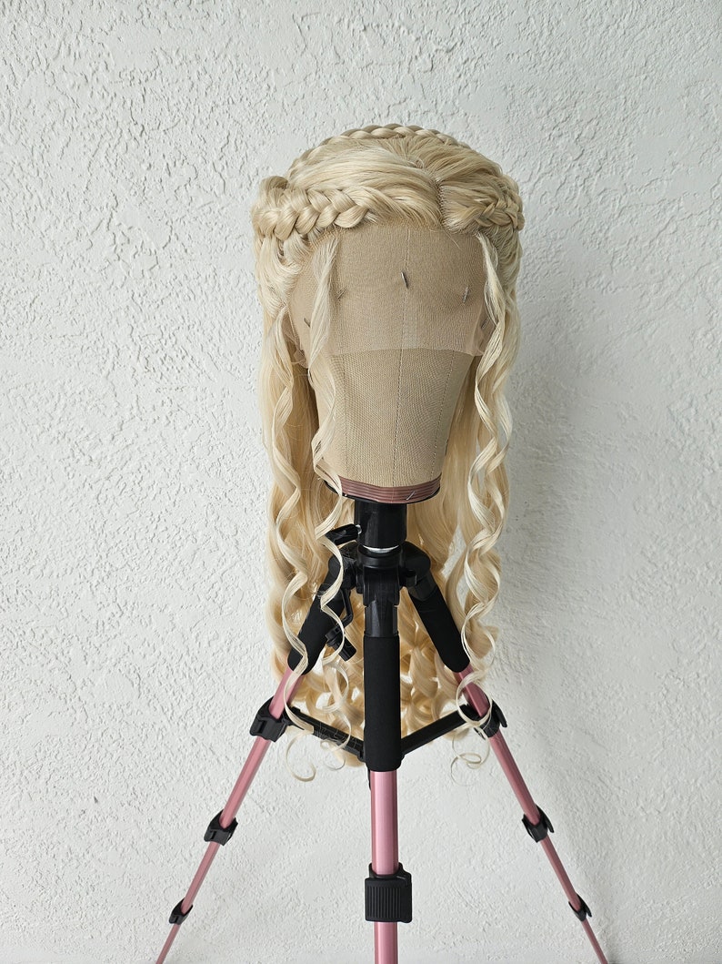 May include: A blonde wig with long, loose curls and a braided crown. The wig is displayed on a pink tripod.
