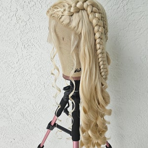 May include: A blonde wig with long, loose curls and two braids styled on the top of the head. The wig is on a mannequin head mounted on a pink tripod.