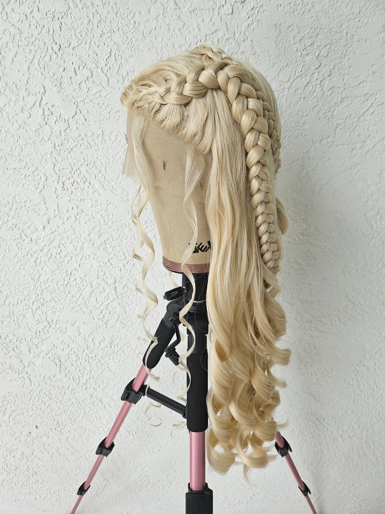 May include: A blonde wig with long, loose curls and two braids styled on the top of the head. The wig is on a mannequin head mounted on a pink tripod.