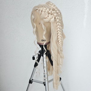 May include: A blonde wig with two braids styled on a mannequin head, mounted on a silver tripod.