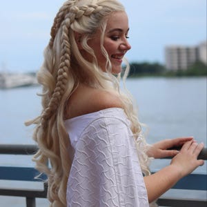 May include: A woman with long, blonde hair styled in braids. She is wearing a white off-the-shoulder dress and is standing near a body of water.