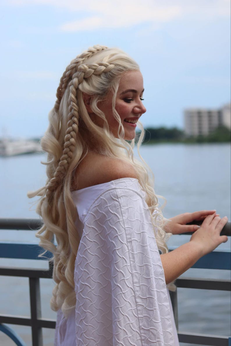 May include: A woman with long, blonde hair styled in braids. She is wearing a white off-the-shoulder dress and is standing near a body of water.