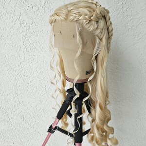 May include: A blonde wig with long, loose curls and a braided crown. The wig is on a wig stand with a pink tripod.