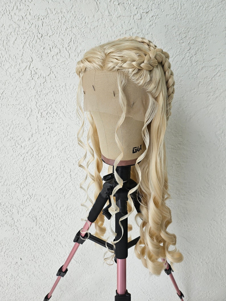 May include: A blonde wig with long, loose curls and a braided crown. The wig is on a wig stand with a pink tripod.
