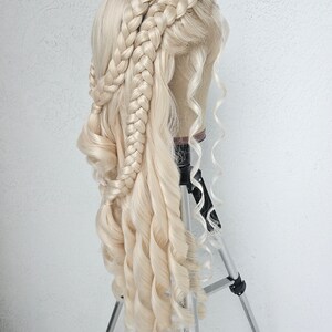 May include: A blonde wig with long, loose curls and three braids styled into a crown. The wig is displayed on a tripod.