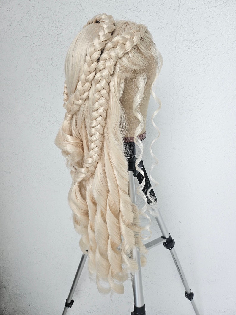 May include: A blonde wig with long, loose curls and three braids styled into a crown. The wig is displayed on a tripod.