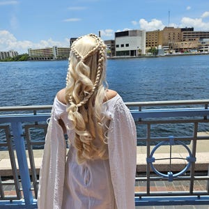 May include: A woman with long blonde hair styled in braids stands facing away from the camera. She is wearing a white dress with a long, flowing cape. The woman is standing on a pier overlooking a body of water. The background shows a city skyline.