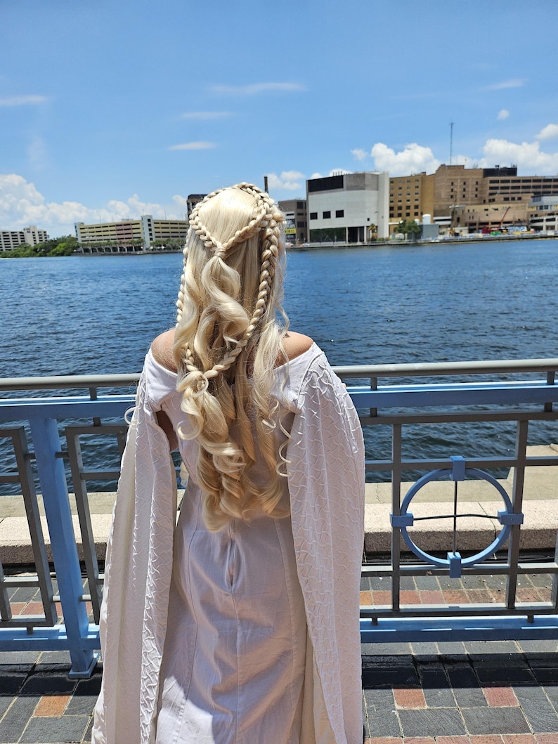 May include: A woman with long blonde hair styled in braids stands facing away from the camera. She is wearing a white dress with a long, flowing cape. The woman is standing on a pier overlooking a body of water. The background shows a city skyline.