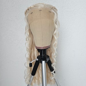 May include: A blonde wig with long, loose curls is displayed on a tripod stand. The wig has a braided crown and is styled in a cascading waterfall of curls.