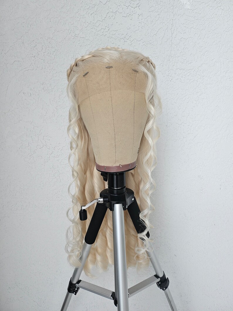 May include: A blonde wig with long, loose curls is displayed on a tripod stand. The wig has a braided crown and is styled in a cascading waterfall of curls.