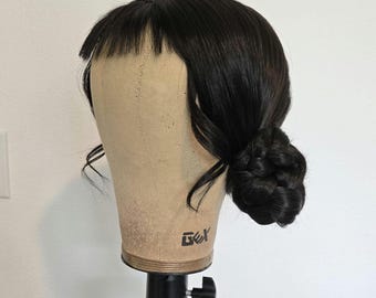 May include: A black wig styled with a side bun and bangs, displayed on a mannequin head. The wig has a natural-looking shine and texture. The mannequin head is mounted on a tripod.