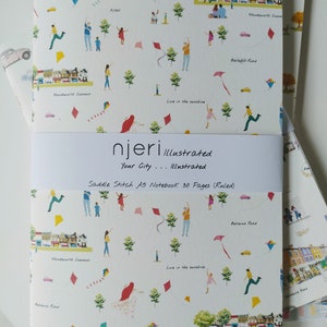 May include: A white notebook with a colourful illustration of kites, people, and houses. The notebook is titled "njeri illustrated" and says "Your City... Illustrated" and "Saddle Stitch A5 Notebook 30 Pages (Ruled)".