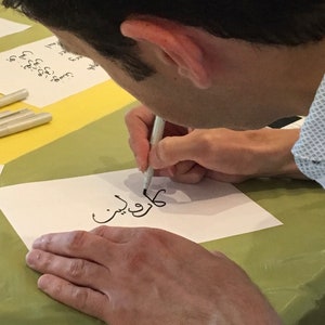 May include: A person writing the Arabic word "كُلّ شيء" in black ink on a white piece of paper with a silver pen.