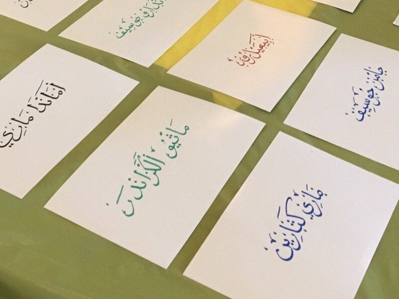 May include: A collection of white cards with Arabic calligraphy in different colors, including blue, green, and red. The text on the cards includes the words "Maatiq Al-Kazand", "Jaman Jaman", "Mary Katerine", "Daroo Josif", and "Amada Mary".