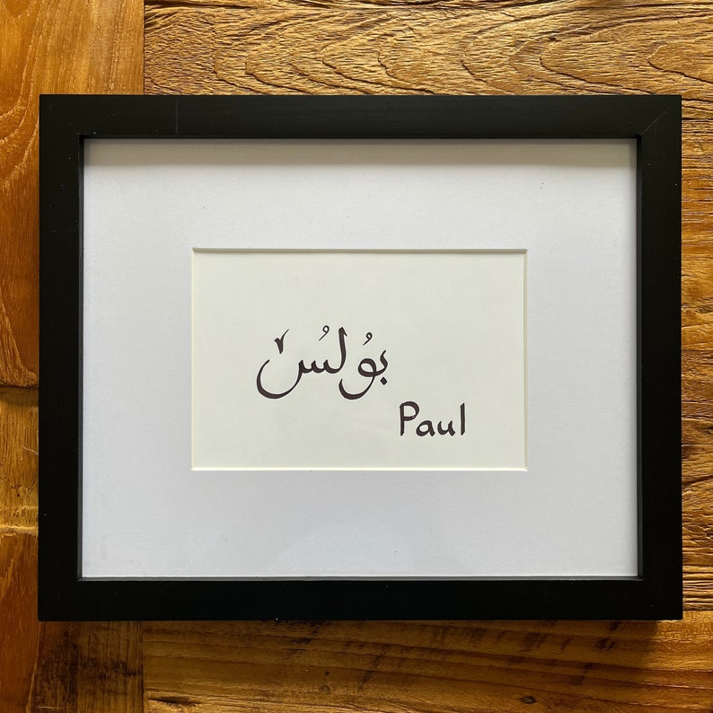 May include: A black framed print with a white background. The print features the Arabic word for "Paul" in calligraphy, followed by the English word "Paul".