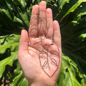 May include: A pair of copper-colored wire earrings in a leaf design. The earrings are held in a hand against a backdrop of green foliage. The earrings have a hook closure.