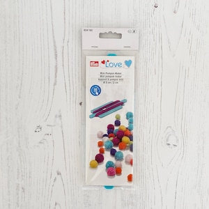 Puede incluir: Un fabricante de pompones de plástico azul y blanco con un diseño en forma de corazón y la palabra "Love" en el embalaje. El embalaje también muestra una variedad de pompones de colores.