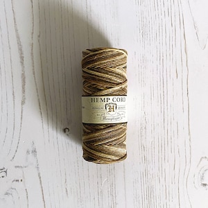 May include: A roll of natural hemp cord in brown, beige and cream colours. The cord is wrapped around a cardboard tube with a label that reads "Hemp Cord, Premium Quality, Est. 1997, Hemptique".