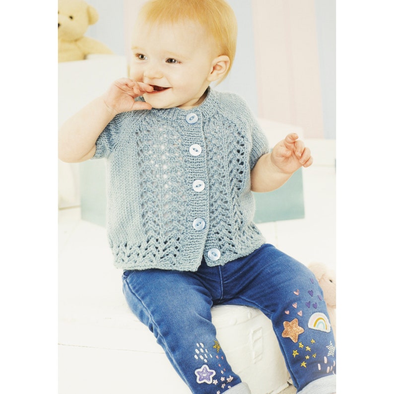 May include: A baby wearing a light blue knitted cardigan with a button closure and blue jeans decorated with colorful patches.