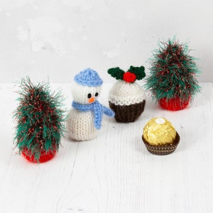 Könnte beinhalten: Ein gestrickter Schneemann, ein gestrickter Weihnachtspudding, ein gestrickter Weihnachtsbaum und eine Schokoladentrüffel in goldener Verpackung. Alle Artikel befinden sich auf einer weißen Oberfläche.