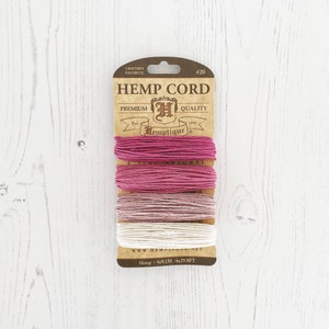 Op de afbeelding: Een verpakking Hemptique henneptouw in drie kleuren: roze, lichtroze en wit. De verpakking is gelabeld met "Hemp Cord" en "Hemptique" met de tekst "4x9,1 m 4x29,8 ft".