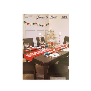May include: A red and white table runner with a snowflake design is set on a dark wood table. The table is decorated for a Christmas dinner with candles, glasses, and crackers. The table is set for four people. The image includes the text "James C. Brett" and "JB812 Intermediate Knit".