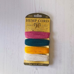 May include: A pack of four spools of hemp cord in pink, teal, yellow, and white. The pack is labelled "Hemp Cord" and "Homatique" with the text "Premium Quality" and "Est. 1997". The pack also states "Hemp 4x9.1M 4x29.8FT".