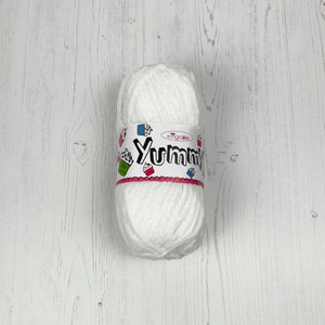 May include: A white ball of yarn with a label displaying the word "Yummy" in a whimsical font. The label is decorated with cupcake illustrations in green, red, and blue. The yarn has a soft, fluffy texture, ideal for crafting.