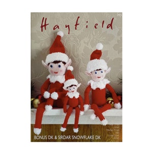Op de afbeelding: Breipatroon voor drie kerstkabouters, een kleine, een middelgrote en een grote kabouter. Het patroon heet "Hayfield" en is bedoeld voor gebruik met Bonus DK en Sirdar Snowflake DK garen. De kabouters dragen rode en witte gebreide mutsen en truien met witte bontkraag. De kabouters zitten op een plank met een kerstboom op de achtergrond.