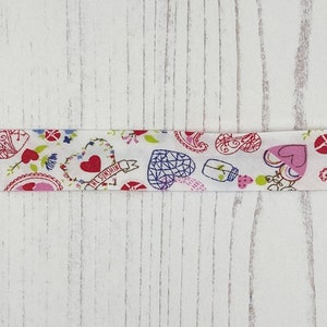 Peut inclure: Ruban en tissu blanc avec un motif rose, rouge et vert de cœurs, de fleurs et de motifs paisley. Le ruban porte l'inscription "The Sunshine".