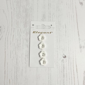 May include: Four white flower-shaped buttons with two holes each, packaged on a white card with the word "Elegant" printed in gold lettering.