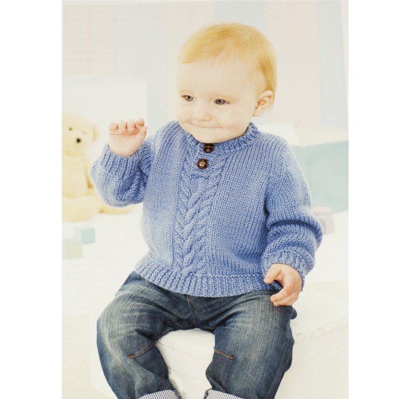 May include: A baby wearing a light blue knitted sweater with a cable knit pattern and a single button closure at the neck. The sweater has long sleeves and is worn over a pair of blue jeans.