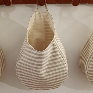Pode incluir: Três cestos de armazenamento suspensos em tecido com um padrão de riscas horizontais bege e branco. Cada cesto tem um corpo arredondado em forma de gota com uma alça de tecido no topo para pendurar num suporte de parede de madeira.
