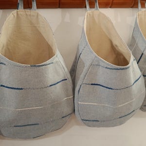 Può includere: Diverse borse portaoggetti sospese con esterno in tessuto azzurro e interno color naturale. Le borse hanno una forma arrotondata e sono decorate con strisce orizzontali blu e bianche. Sono sospese a un'asta di legno.