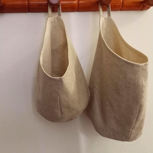 Könnte beinhalten: Zwei beigefarbene Hängetaschen aus Leinen mit schlichtem Design. Die Taschen hängen an einer Holzstange.