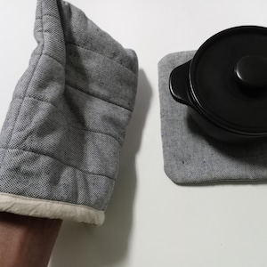 Pode incluir: Uma luva de forno cinza acolchoada com acabamento branco e um descanso de panela quadrado cinza correspondente. Uma panela preta com tampa repousa sobre o descanso de panela. Acessórios de cozinha e jantar.