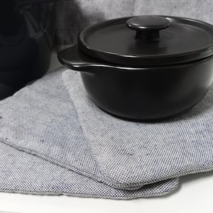 Pode incluir: Uma caçarola de cerâmica preta com tampa, repousando sobre uma pilha de panos de prato de tecido cinza e branco estampados. A tigela tem formato arredondado com uma alça em cada lado. A tampa tem um pequeno botão redondo.